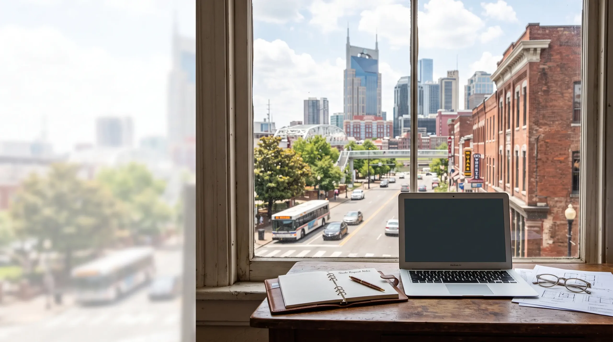 An open notebook and laptop rest on a wooden desk by a window with a view of a city street and skyline.
